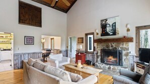 A spacious living room with a stone fireplace, a dining area, and a kitchen visible in the background. - Hidden Birch at Sugarbush (Fayston)