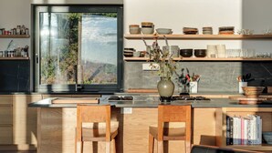 Eine moderne Küche mit einem großen Fenster, hölzernen Barhocker und einer Auswahl an Geschirr und Besteck auf Regalen.