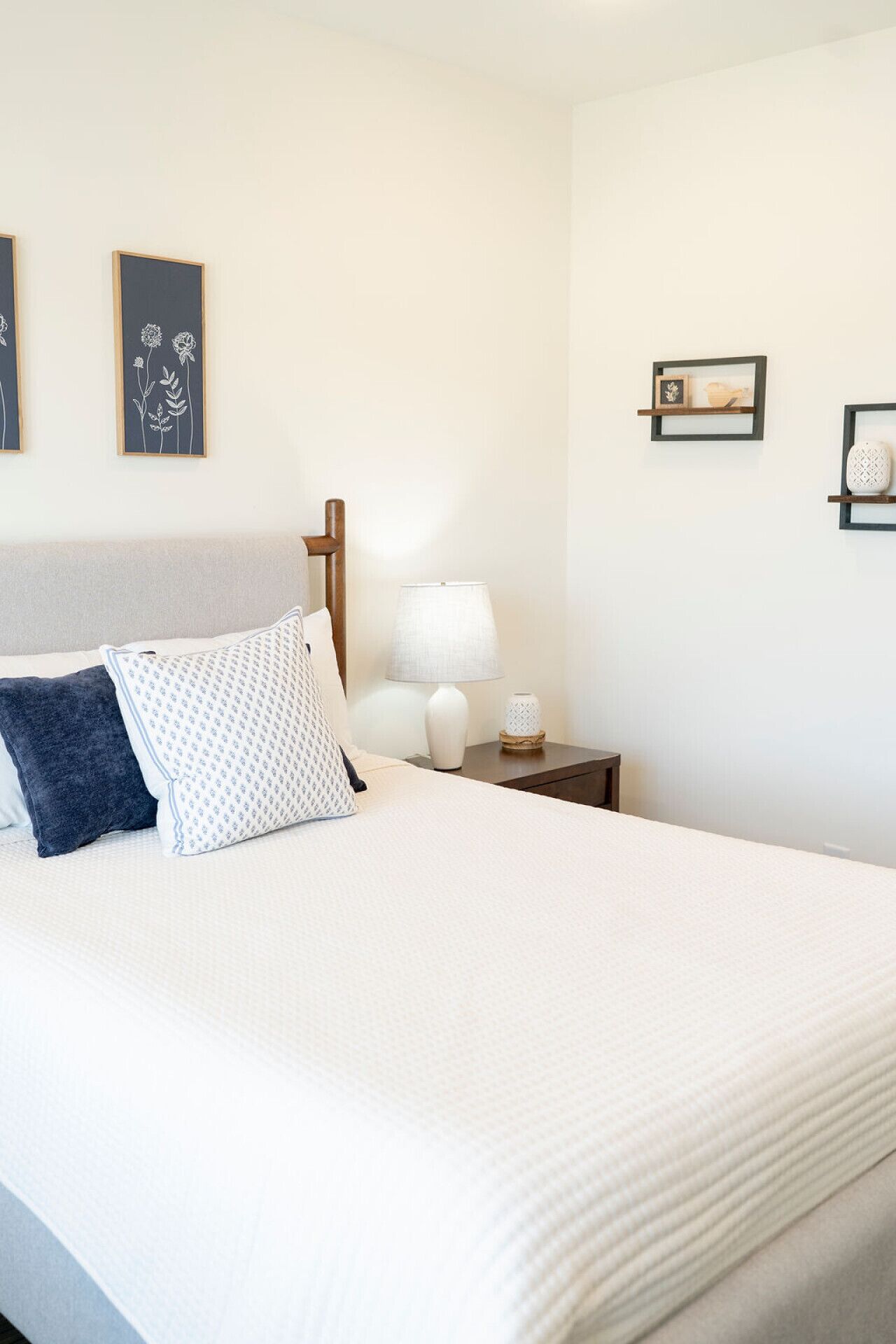 A neatly made bed with white bedding and two pillows, a bedside lamp, a small table with a vase, and two wall-mounted shelves with decorative items.