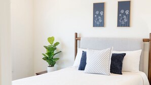 Ett sovrum med en säng, en takfläkt, två inramade konstverk med blommotiv, en krukväxt och en mörk matta.
