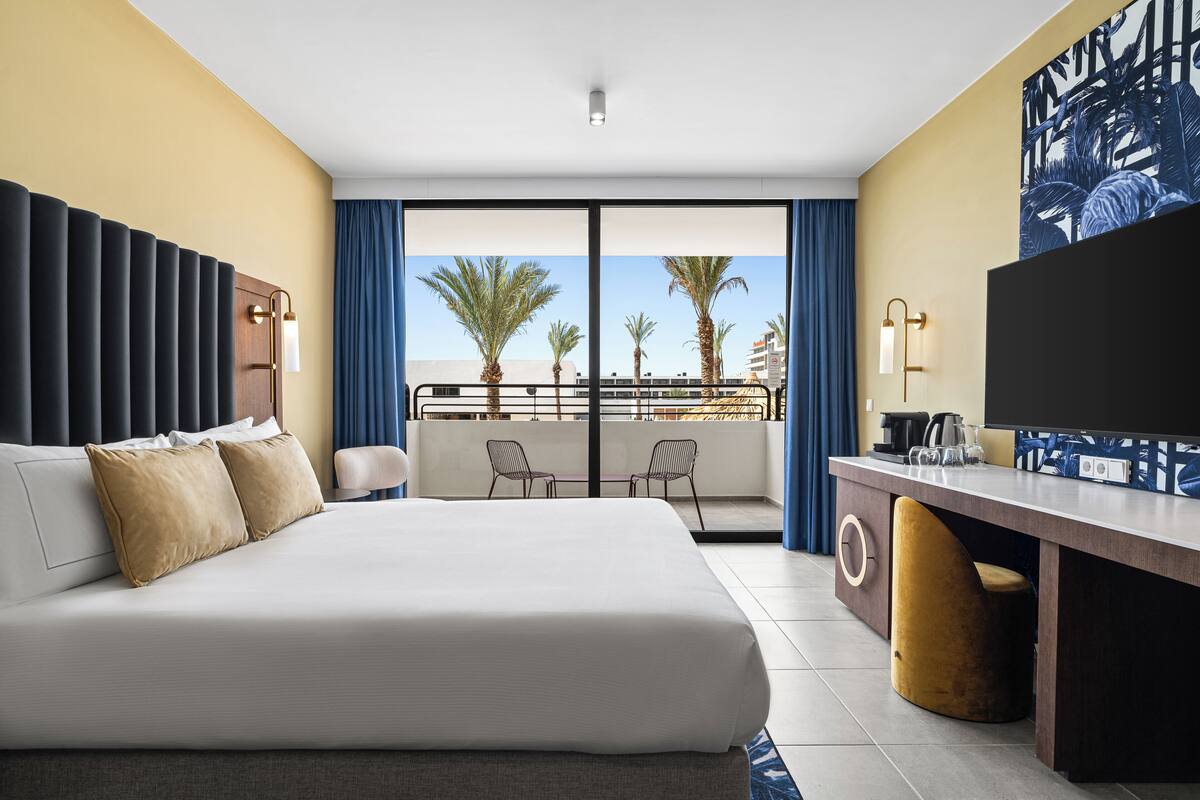 A hotel room with a large bed, a desk with a chair, a TV, and a balcony with palm trees and a view of the city.