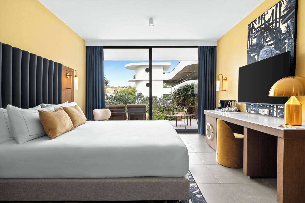 A modern hotel room with a large bed, a desk, and a view of the cityscape through the window.