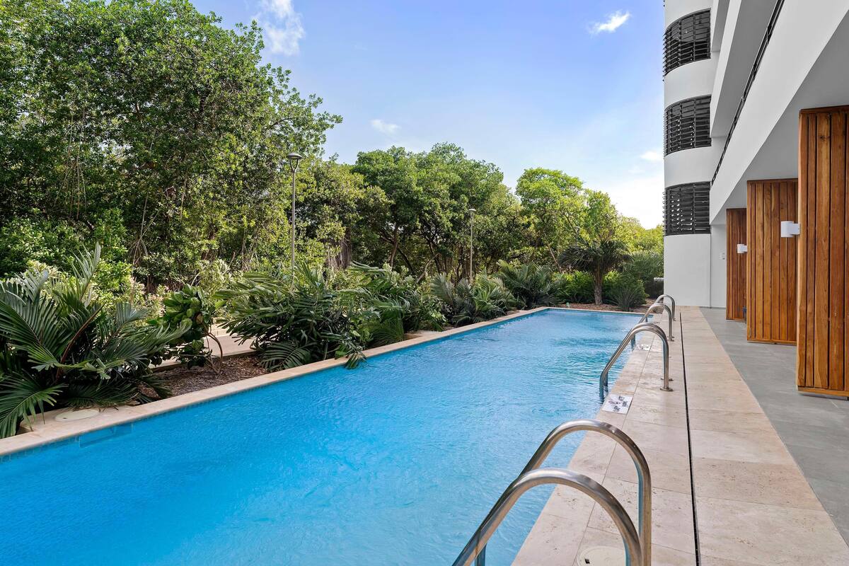 A swimming pool with metal railings, surrounded by lush greenery and a modern building with wooden doors.