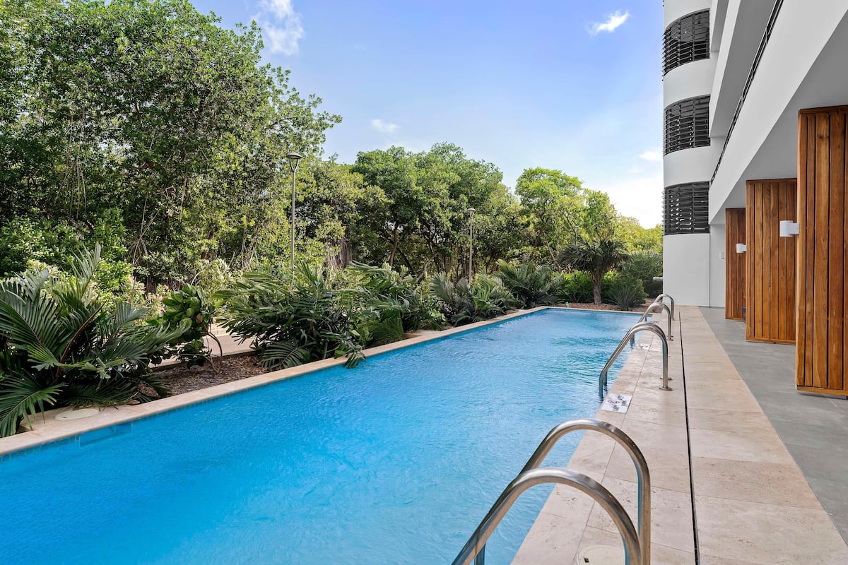 Una piscina con barandillas metálicas, rodeada de exuberante vegetación y un edificio moderno con puertas de madera.