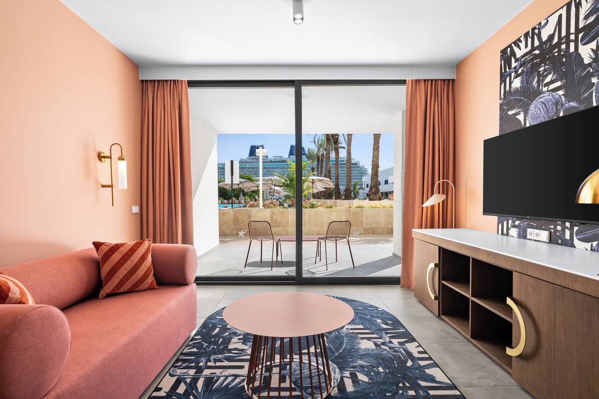 A modern hotel room with a sofa, a round table, a TV, and a balcony with a city view.