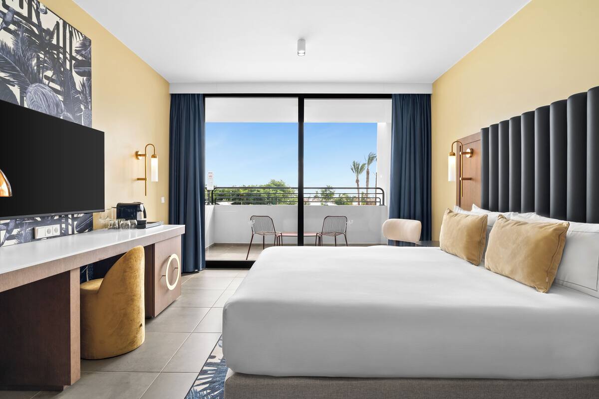 A hotel room with a large bed, a desk with a flat-screen TV, a balcony with chairs, and a view of palm trees.
