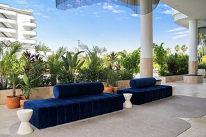 A modern outdoor seating area with blue sofas and white side tables, surrounded by potted plants and a clear view of a white building in the background. - The Rif at Mangrove Beach Corendon All-inclusive (Willemstad)