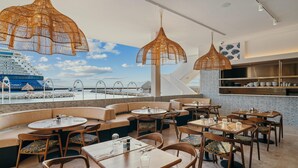 Un restaurante con mesas redondas y cabinas, con una gran embarcación visible a través de las ventanas, y lámparas colgantes de mimbre.