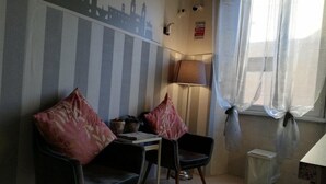 Una habitación pequeña y acogedora con papel tapiz a rayas, dos sillas con cojines rosa, una mesa, una lámpara y una ventana con cortinas traslúcidas.