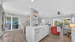 A spacious kitchen with a large island, a dining area with a glass door leading to a balcony, and a living area with a red ottoman and a sofa.