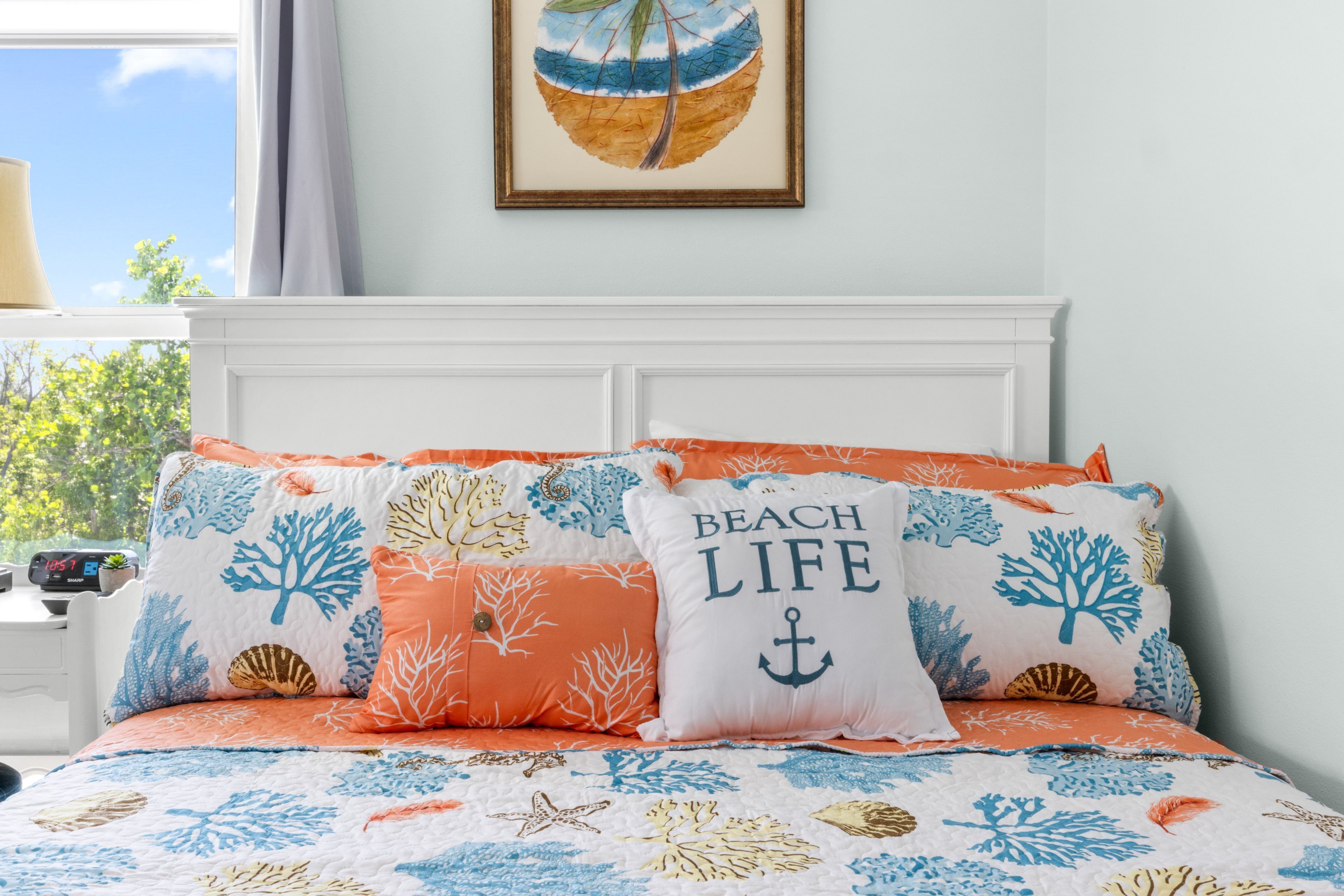 A bed with a beach-themed bedspread and pillows, a framed picture on the wall, and a window with curtains.