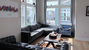 Un salon moderne avec un canapé d’angle, un fauteuil bleu et une table basse agrémentée de bougies. De grandes fenêtres offrent une vue sur le quartier.