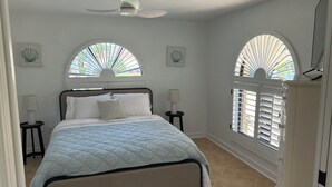 A bedroom with a bed, two bedside tables, a ceiling fan, and two windows with shutters. - 3-bedroom house in fantastic Sanibel with AC (Sanibel)