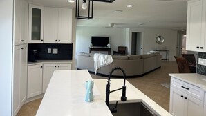 A modern kitchen with white cabinetry, a central island with a sink, and a dining area with a sofa and TV. - 3-bedroom house in fantastic Sanibel with AC (Sanibel)