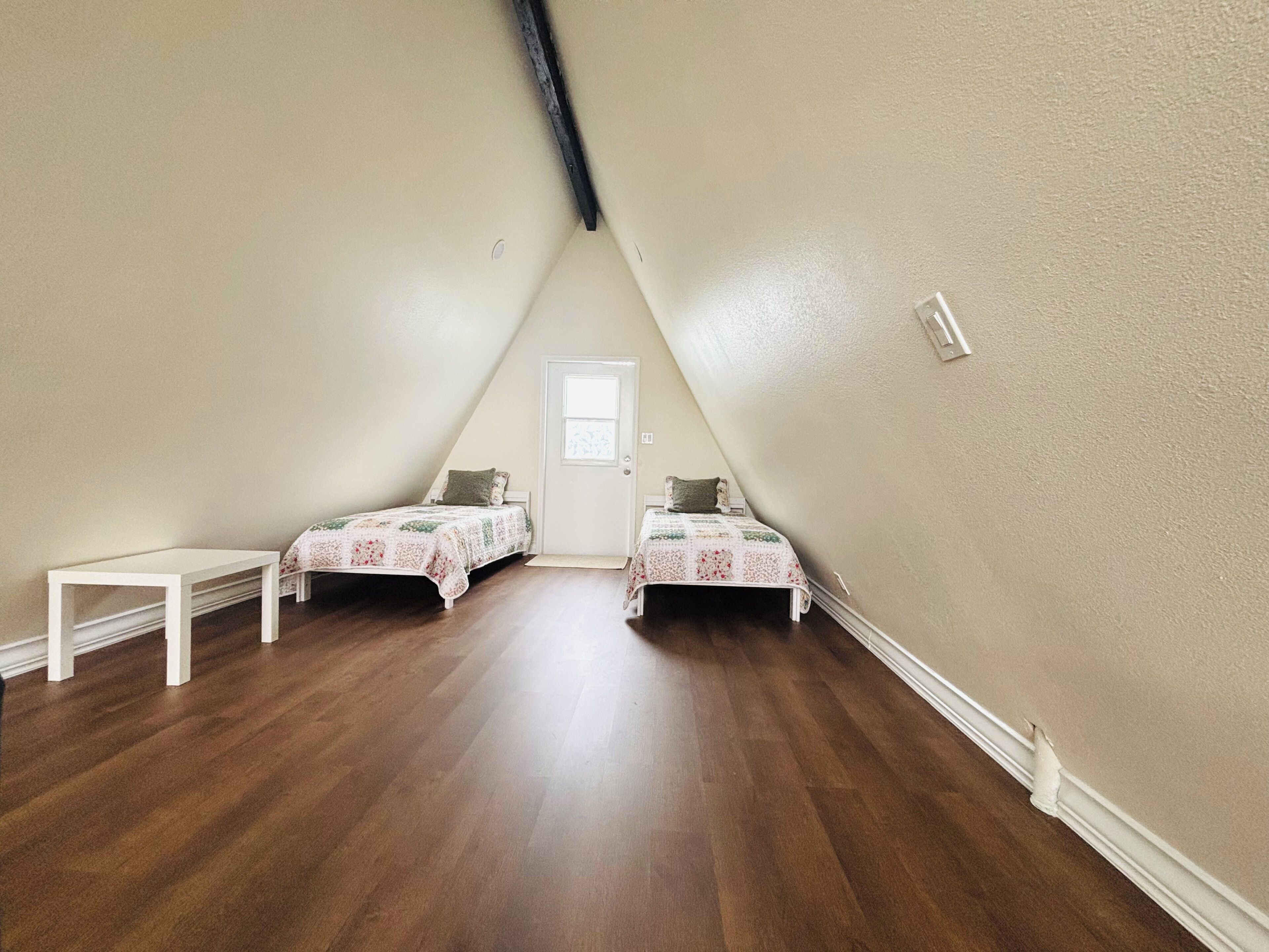 Une petite chambre mansardée triangulaire, avec deux lits, un banc blanc et une porte.