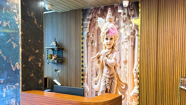 A reception area with a wooden reception desk, a wall-mounted shelf with decorative items, and a mural of a deity. - Hotel Om Palace (Vrindavan)