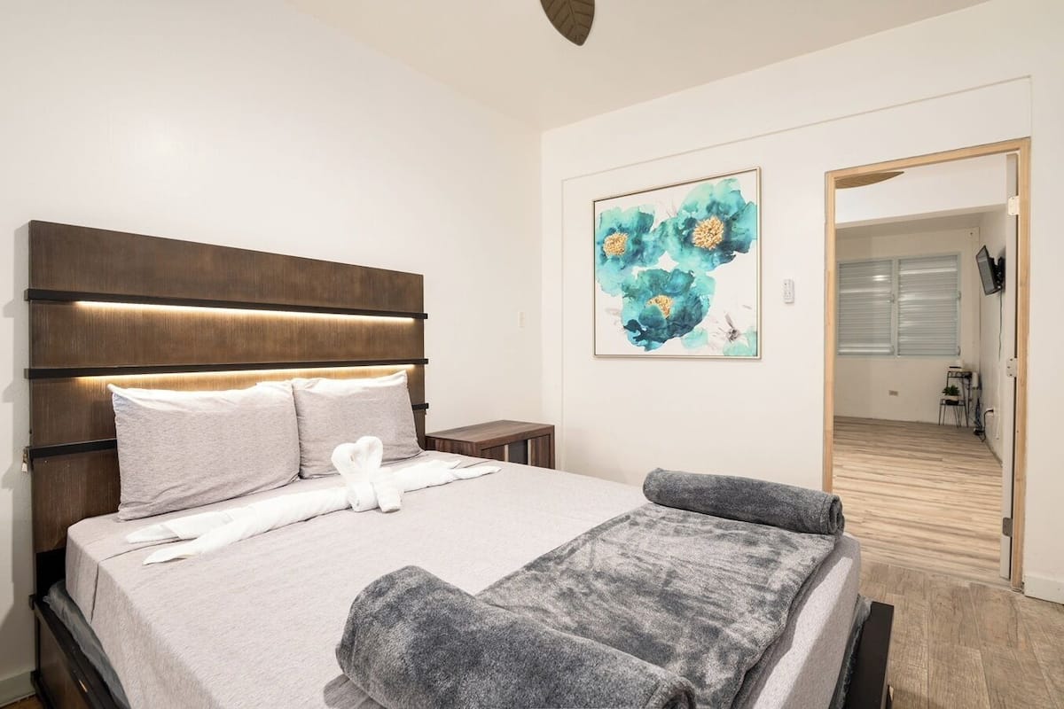A modern hotel room with a large bed, a headboard with built-in lighting, a framed abstract painting, and a view into the hallway.