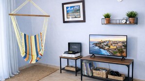 A room with a hammock, a framed picture, a television, and shelves with plants and a clock. - RainForest House (Ceiba)