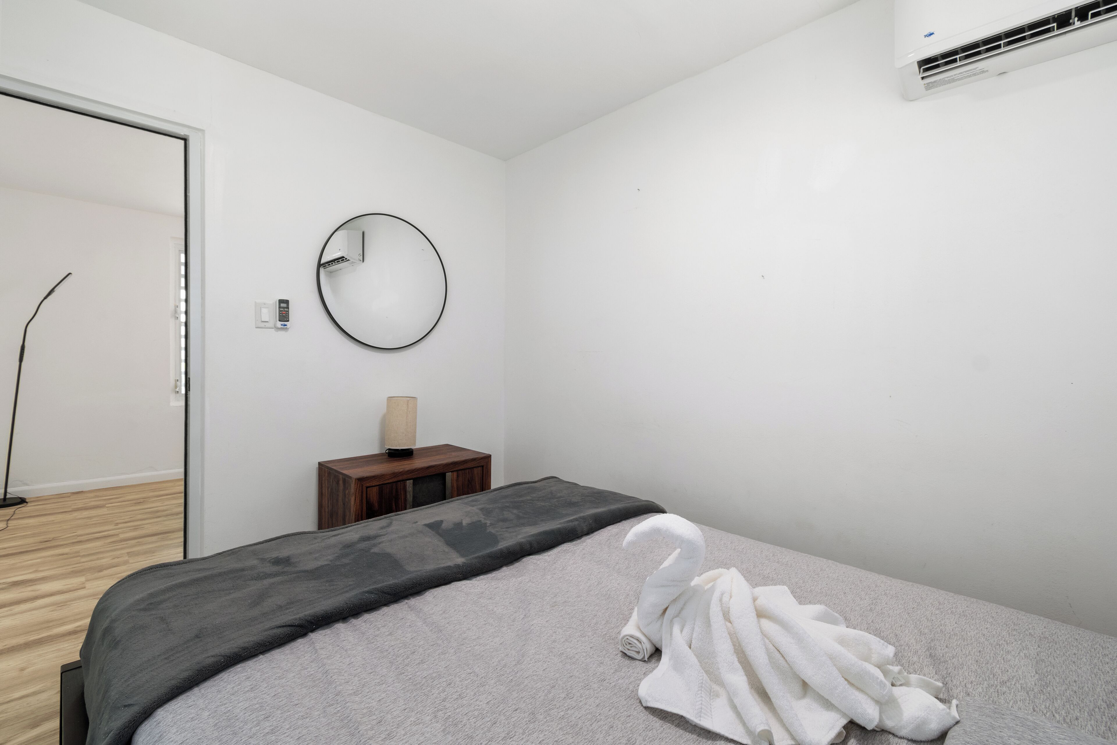 Ein schlicht eingerichtetes Schlafzimmer mit Bett, rundem Spiegel, kleinem Nachttisch und Klimaanlage.