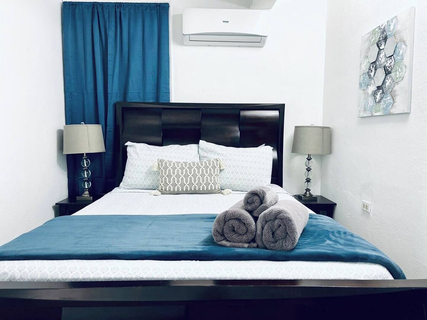 A hotel room with a bed, two bedside lamps, a blue curtain, a wall-mounted air conditioner, and a piece of abstract art.