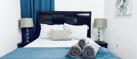 A hotel room with a bed, two bedside lamps, a blue curtain, a wall-mounted air conditioner, and a piece of abstract art.