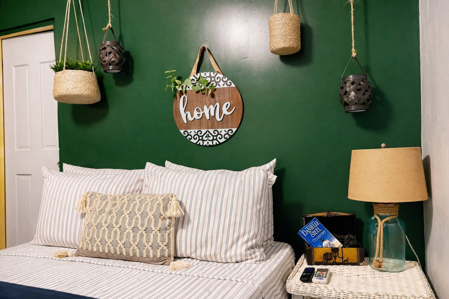 Quarto com parede verde, roupa de cama listrada, planta pendente, placa com a palavra 'home', luminária e mesa de cabeceira com um livro e um celular.