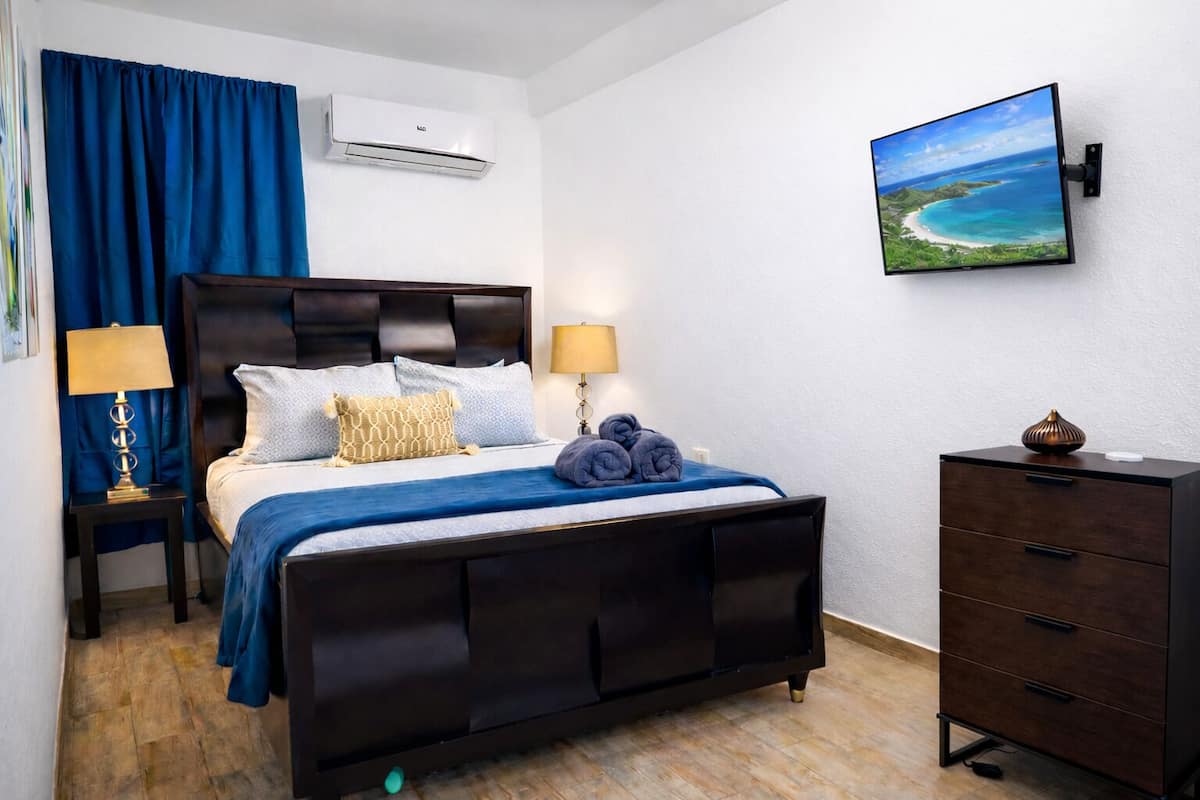 A hotel room with a bed, bedside lamps, a dresser, an air conditioning unit, and a wall-mounted TV.