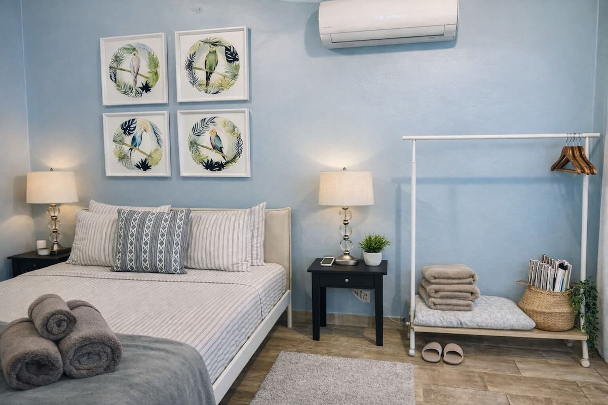 A bedroom with a bed, two bedside tables, a wall-mounted air conditioner, a bench with towels, and framed artwork on the wall.
