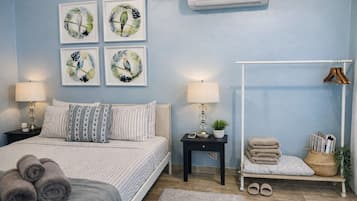 Un dormitorio con cama, dos mesitas de noche, aire acondicionado de pared, un banco con toallas y cuadros en la pared.