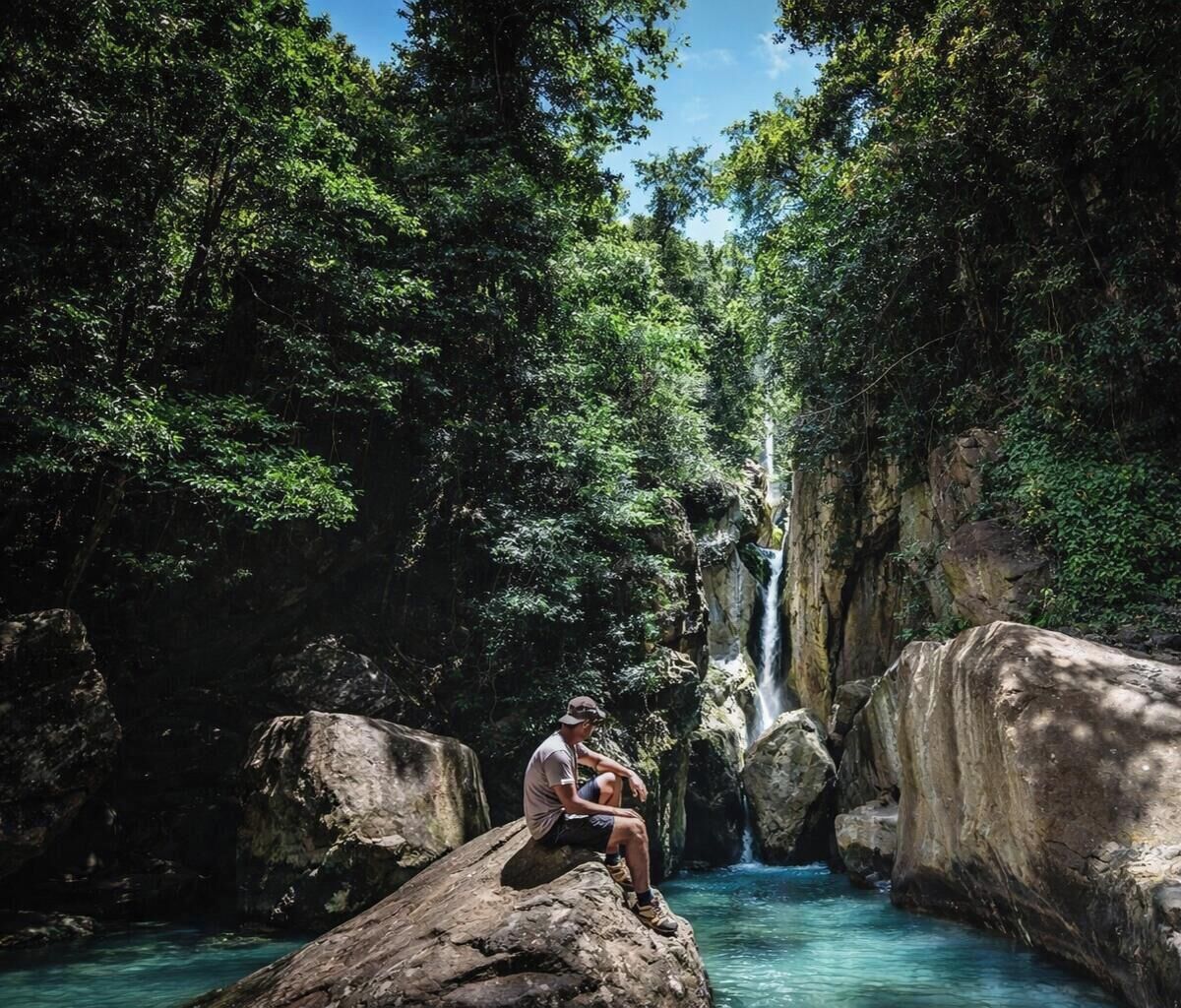 Henkilö istuu kiven päällä vesiputouksen lähellä, metsäisessä ympäristössä.