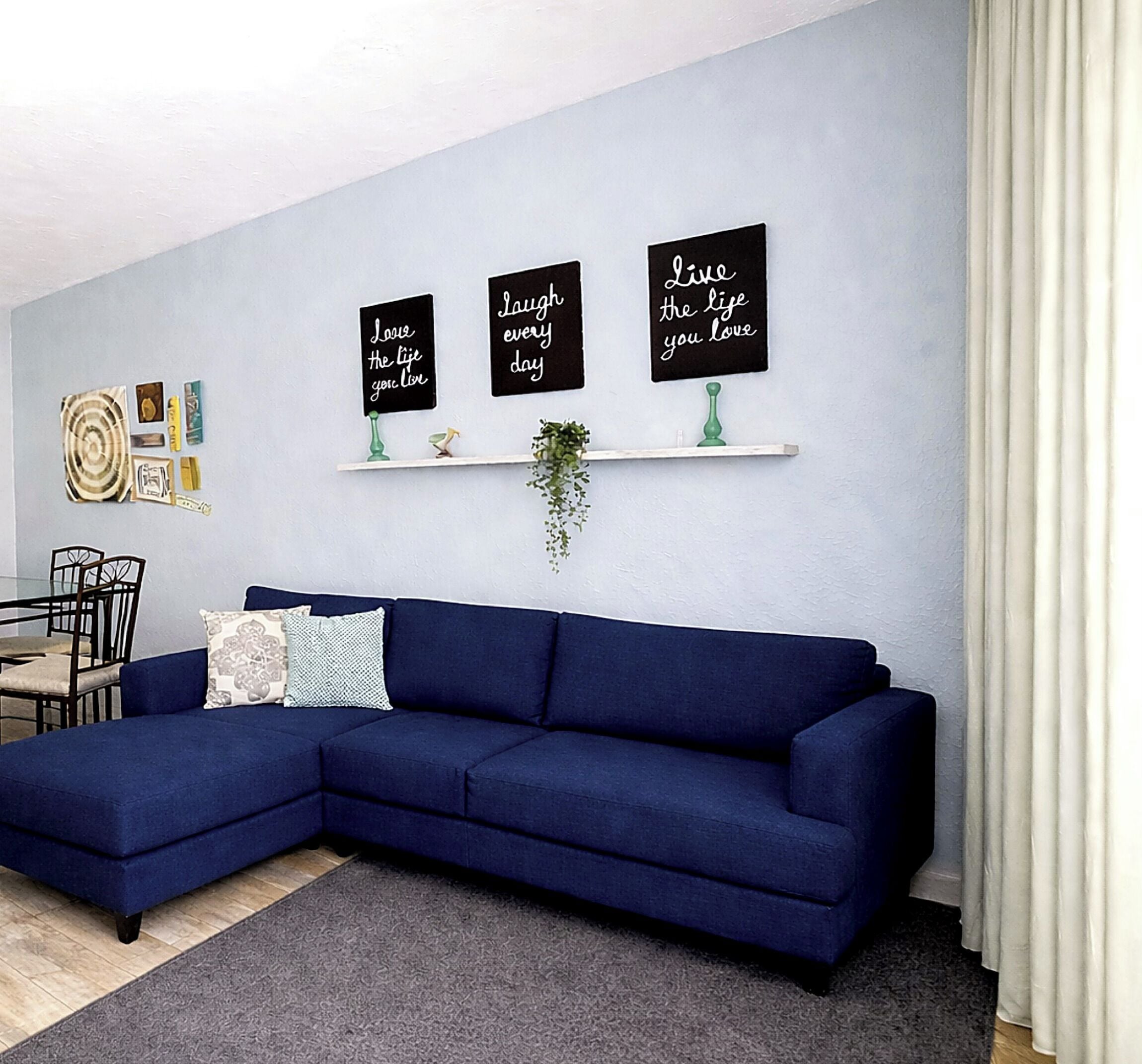 A living room with a blue sectional sofa, a small dining table, and three wall-mounted signs with positive messages.
