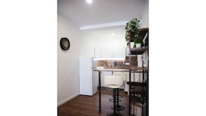 A compact kitchen with white cabinets, a dining area with a white chair, and a black clock on the wall.