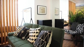 A green sofa with patterned cushions, a black chair, and a wooden partition in a well-lit room with framed pictures on the wall.
