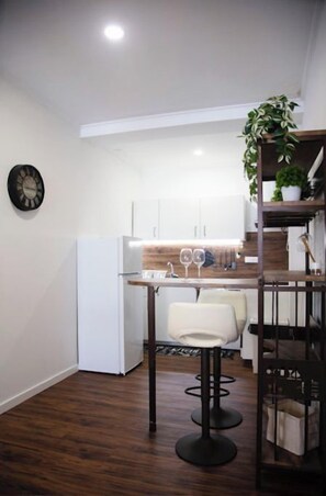 A compact kitchen with white cabinets, a dining area with a white chair, and a black clock on the wall. - Cristal apartamento 1º drt praça velha (Angra do Heroísmo)