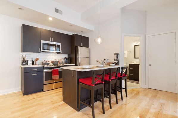 A modern kitchen with dark cabinets, a microwave, stove, and refrigerator. There is a bar area with red cushioned chairs.