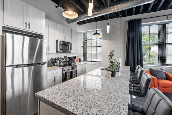 Cocina moderna con electrodomésticos de acero inoxidable, encimera de granito y una zona de estar con sillones de cuero y una planta.