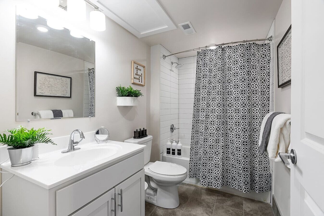 Une salle de bain moderne avec un lavabo blanc, un miroir et des toilettes. On y trouve un rideau de douche à motifs géométriques et une petite plante posée sur le comptoir.