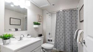 Un baño moderno con un lavabo blanco, espejo y retrete. Hay una cortina de ducha con un estampado geométrico y una planta pequeña en el mostrador.