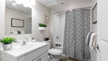 Une salle de bain moderne avec un lavabo blanc, un miroir et des toilettes. On y trouve un rideau de douche à motifs géométriques et une petite plante posée sur le comptoir.