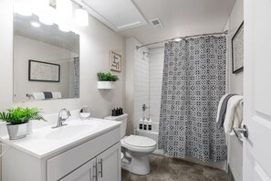 A modern bathroom with a white sink, mirror, and toilet. There is a shower curtain with a geometric pattern, and a small plant on the counter.