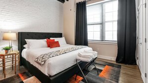 Una habitación de hotel moderna con cabecera negra, ropa de cama blanca y vistas de la ciudad a través de amplias ventanas.