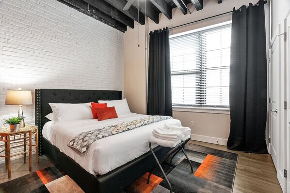 A modern hotel room with a black headboard, white bedding, and a view of the city through large windows.