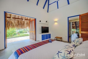 A modern bedroom with a large bed, a TV on a blue cabinet, and a view of a garden through an open door. - New KeyPöint: Tröpical House with incredible view! (Palomino)