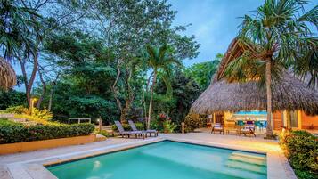 A swimming pool surrounded by lounge chairs and a thatched-roof structure, likely a bar or dining area, set in a lush garden.