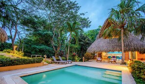 A swimming pool surrounded by lounge chairs and a thatched-roof structure, likely a bar or dining area, set in a lush garden. - New KeyPöint: Tröpical House with incredible view! (Palomino)