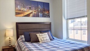 A bedroom with a cityscape view, a bed with plaid bedding, and a nightstand with a lamp.