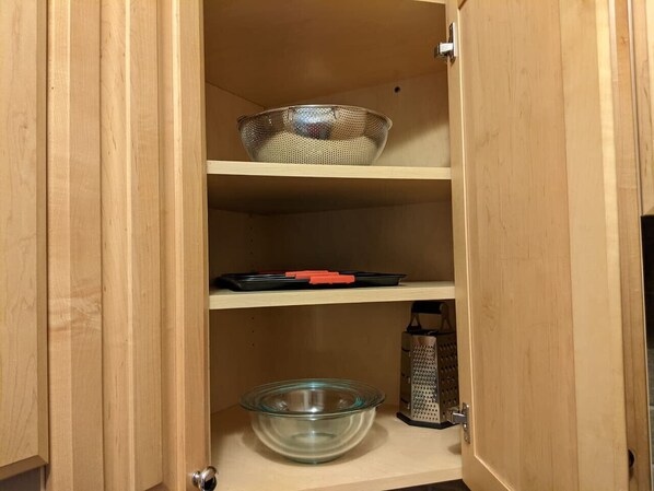Un mueble de madera con tres estantes; el estante superior sostiene un colador de metal, el estante central tiene una bandeja negra con asas rojas, y el estante inferior contiene un tazón de vidrio y un rallador de metal.