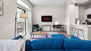 Een moderne woonkamer met een blauwe bank, een televisie waarop 'NETFLIX' wordt weergegeven, en een keuken met witte kasten en een magnetron.