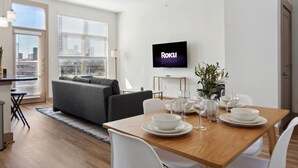 A modern living room with a dining area, a grey sofa, a TV on the wall, and a city view through the windows. - Stylish 1Br in Deep Ellum with Free Parking Near Baylor Med (Dallas)