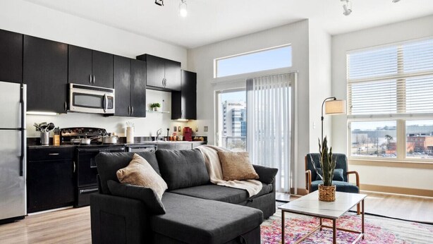 Un apartamento moderno con cocina, un sofá seccional gris, una pequeña mesa de comedor y un ventanal con vista a la ciudad.